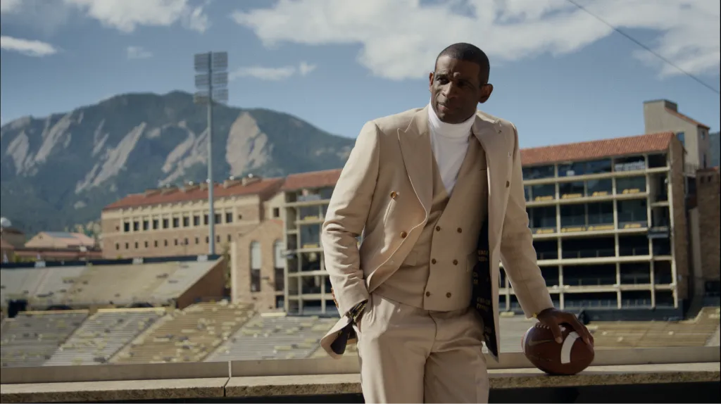 Man in a tan double-breasted suit holding a football in a stadium