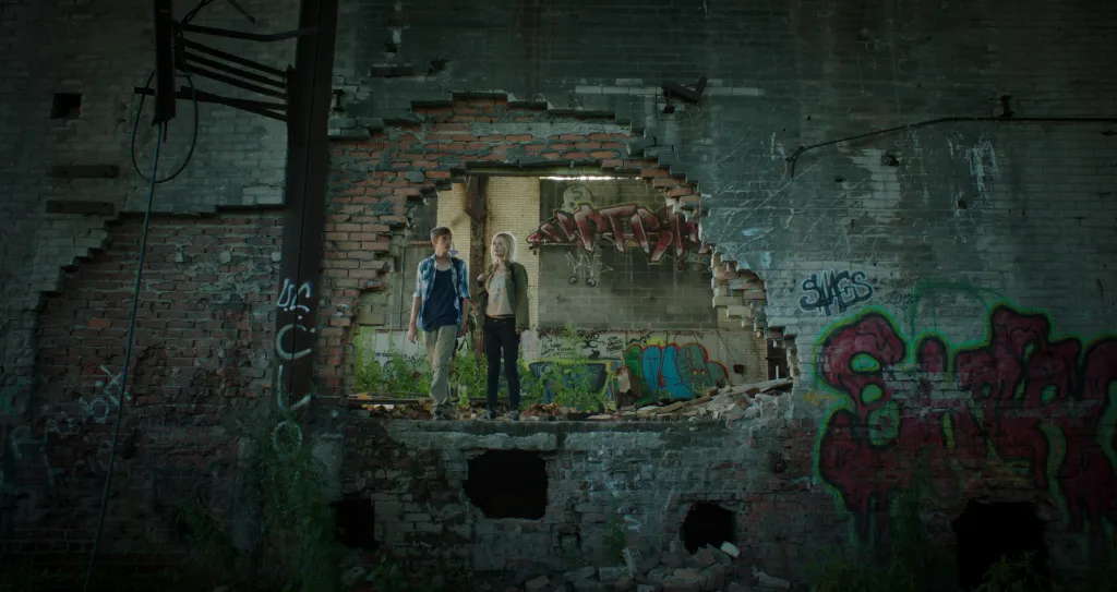 Two figures framed by a broken brick wall in a derelict building